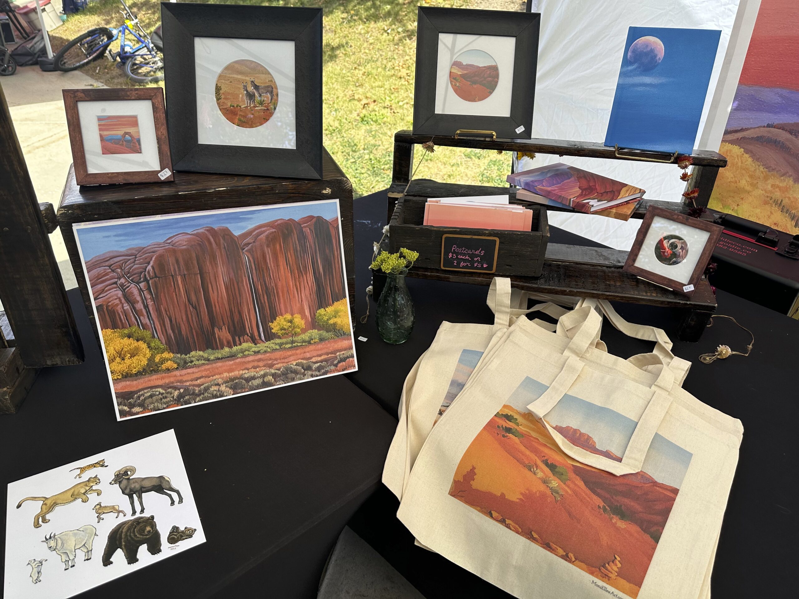 Photo of artwork at farmers market, a painted tote bag, framed oil paintings of deserts small and large, and painted journals.